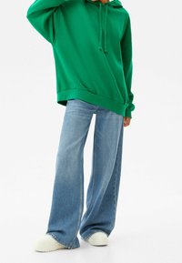 Sudadera verde oversize con capucha, combinada con jeans de pierna ancha de color azul claro y zapatillas blancas. Tejido casual y dise�ño de corte relajado.