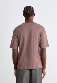 Tricou din bumbac maro cu mâneci scurte, guler rotund, textură netedă și croială lejeră, văzut din spate.