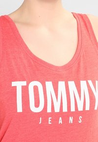 Röd linne tillverkat av mjukt tyg med stora vita "TOMMY" bokstäver och mindre "JEANS" text nedanför, med rundad halsringning.