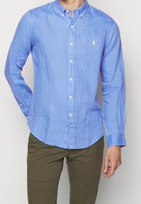 Light blue button-up shirt in linen fabric with long sleeves, a button-down collar, and a small yellow logo on the chest.