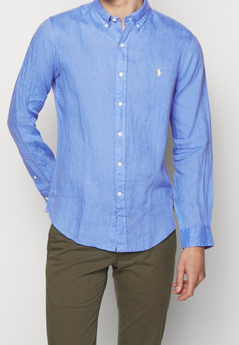 Light blue button-up shirt in linen fabric with long sleeves, a button-down collar, and a small yellow logo on the chest.