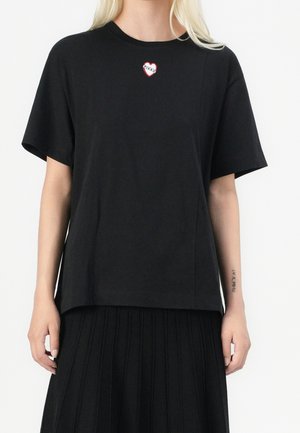 Vrouw draagt een zwart T-shirt met korte mouwen met een klein rood hartje en het "Pinko" logo op de borst, gecombineerd met een zwarte geplooide rok.