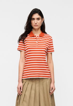 Femeie cu păr lung și închis la culoare, purtând un tricou polo cu dungi portocalii și albe și o fustă plisată bej, stând în fața unui fundal simplu.