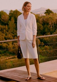 Femme portant un blazer blanc et un short arrivant aux genoux, tenant un sac à main noir, debout sur une terrasse en bois avec un arrière-plan de verdure.