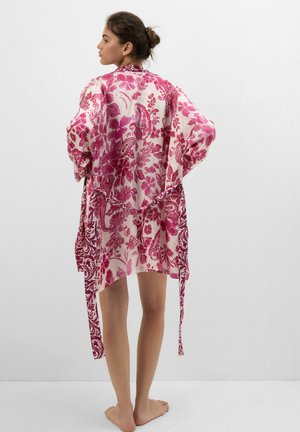 Vrouw staat blootsvoets met de rug naar ons toe, draagt een losse, lange mouwen roze-witte bloemenpatroon badjas met hangende stoffen stroken.