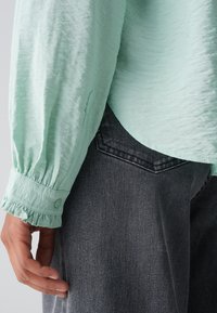 Blusa verde menta, con texture, maniche a bufalo e polsini a balze, infilata in jeans di denim grigio scuro con una cucitura visibile della tasca posteriore.