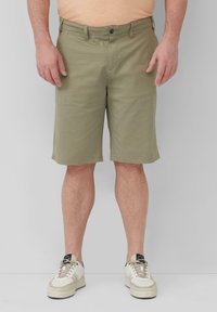 Khaki-Shorts mit flacher Vorderseite, Gürtelschlaufen und einem einzigen Knopfverschluss. Getragen mit weißen Sneakers mit beigen Akzenten und Schnürsenkeln.