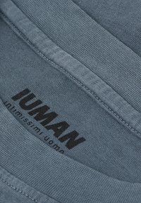 Niebieska bawełniana tkanina o ribbowej strukturze z czarnym nadrukowanym logo. Posiada gładki dekolt i widoczne szwy na brzegach.