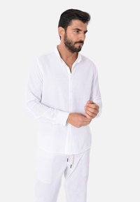Camicia bianca in lino con maniche lunghe e colletto rilassato, dotata di una chiusura con bottoni sul davanti e una finitura leggera e testurizzata.