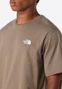 Lähellä otettu kuva henkilöstä, joka pitää päällään ruskeaa lyhythihaista t-paitaa, jossa on valkoinen The North Face -logo painettuna rintaan.