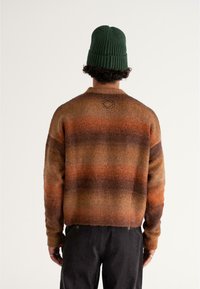 Gestreifter Pullover in Braun und Orange, aus strukturiertem Strickmaterial, mit Rundhalsausschnitt und gesticktem Logo auf der Rückseite. Kombiniert mit dunklen Hosen.