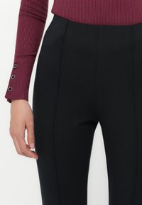 Donna che indossa pantaloni neri a vita alta e attillati e un top a maniche lunghe color bordeaux a costine con tre bottoni neri sui polsini.