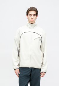 Ανοιχτό μπεζ φλις ζακέτα με φερμουάρ και ψηλό γιακά, με αντίθετους σκούρους γκρίζους τόνους και τσέπη στο μπροστινό στήθος που φέρει το λογότυπο της Adidas.