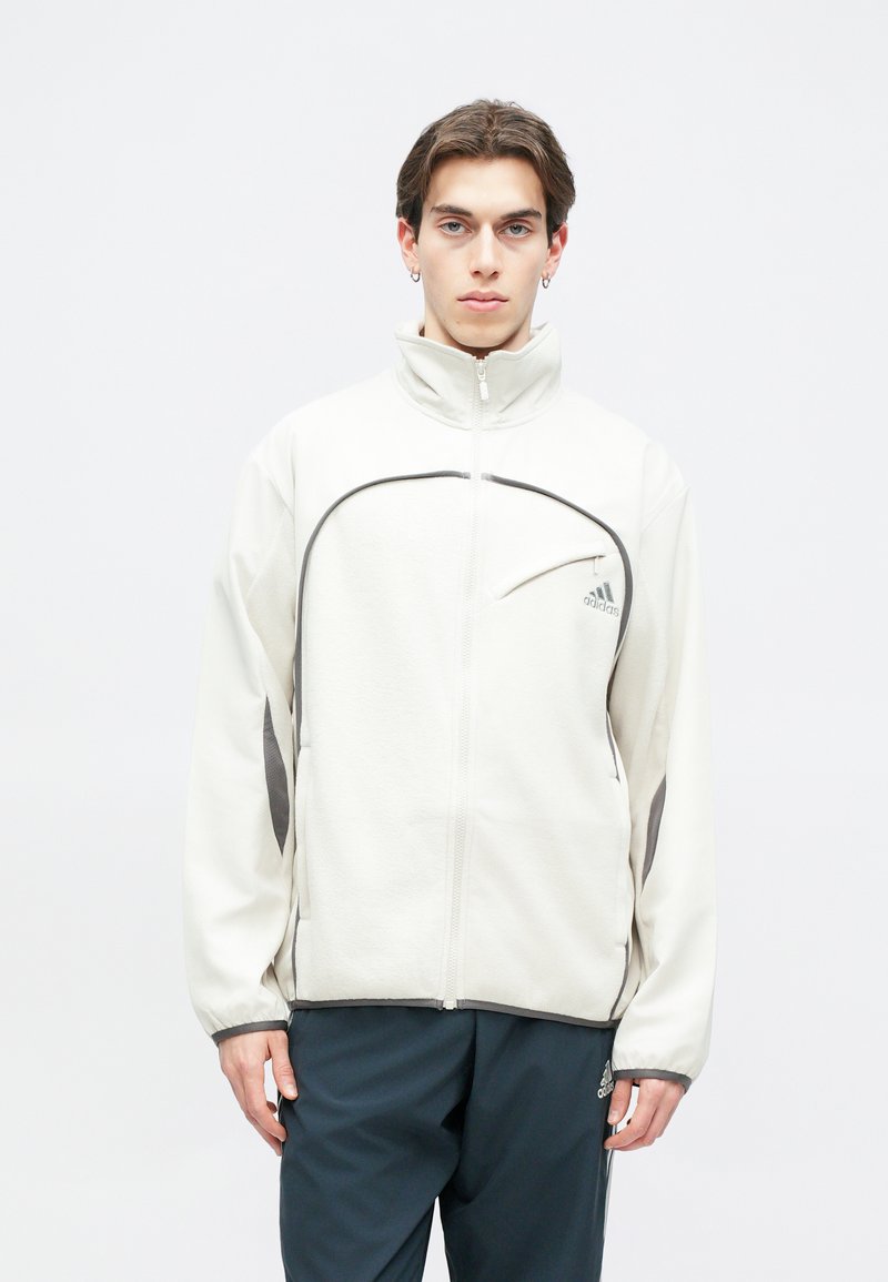 Lichtbeige ritjack met hoge kraag, contrasterende donkergrijze accenten en een voorzak op de borst met het Adidas-logo.