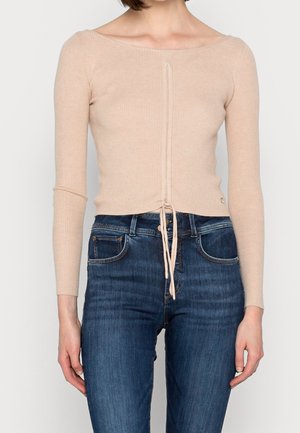 Helle beigen, geribbeltes langärmliges Oberteil mit rundem Ausschnitt, mit Kordelzug am Saum, getragen mit dunkelblauen Jeans.