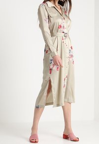 Abito camicia midi color khaki chiaro con stampa floreale, maniche lunghe, vita con cintura e spacchi laterali. Indossato con sandali rosa senza chiusura.