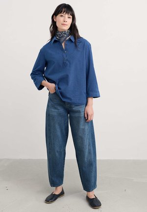 Blaues, gestreiftes Hemd mit Knopfleiste und dreiviertel Ärmeln, kombiniert mit lockeren blauen Jeans und schwarzen Slippern. Dezenter Kragen.