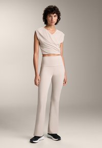 Bež kratki top s V izrezom i drapiranim dizajnom, u kombinaciji s pripijenim hlačama s visokim strukom i flared krojem u odgovarajućoj nijansi. Crne tenisice upotpunjuju odjevnu kombinaciju.