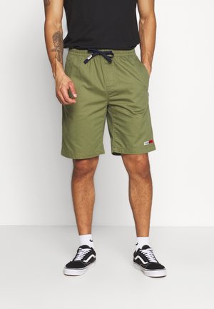Mies pukeutuneena oliivinvihreisiin shortsihin, joissa vetoketjut, mustaan t-paitaan, valkoisiin Nike-sukkiin ja mustiin Vans-tennareihin, seisomassa käsineen taskuissa.