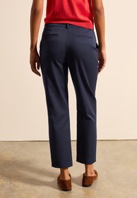 Granatowe spodnie o dopasowanym kroju, z tylnymi kieszeniami i gładką fakturą. Noszone z czerwoną bluzką i brązowymi butami wsuwanymi.