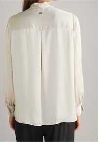 Blusa crema con maniche lunghe, dettaglio a pieghe sul retro, texture satinata e polsini con bottoni. Presenta un piccolo logo metallico sul collo.