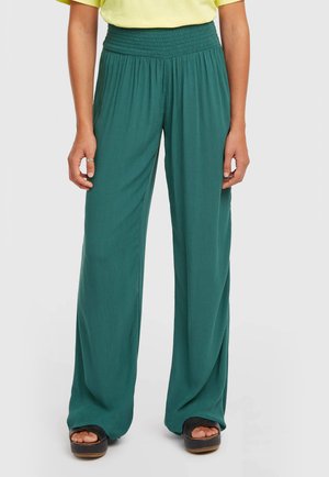 Szmaragdowe spodnie o szerokich nogawkach z marszczoną gumką w pasie, o płynnej fakturze i luźnym kroju. Połączone z ciemnymi sandałami na platformie.
