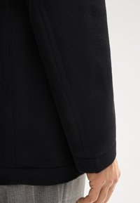 Giacca in pile nera con una texture liscia, caratterizzata da una vestibilità su misura e una manica con cuciture rifinite. Mano visibile che poggia sul tessuto.