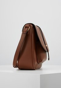 Ruskean teksturoitu nahkainen olkalaukku läpällä ja säädettävällä hihnalla, esillä valkoisella pinnalla yksinkertaisen taustan edessä.