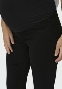 Jeans de maternité noirs au design ajusté, texture lisse et poches latérales, montrant un gros plan de la zone de la taille et du placement des mains.