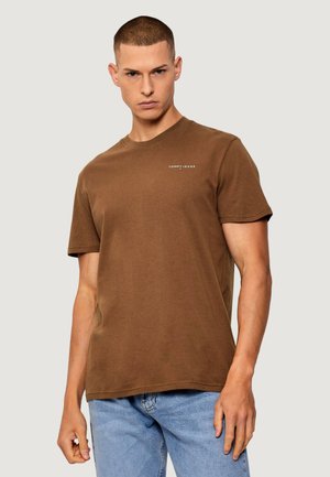 Giovane uomo con capelli corti che indossa una maglietta marrone semplice Tommy Jeans e jeans azzurri, in piedi contro uno sfondo chiaro.