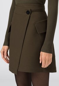 Olivgrön omslagsskjorta tillverkad av slitstarkt tyg, med sidoknappar, kontrasterande fickor och rak fåll.