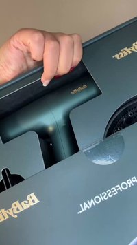 Robust blågrønn hårføner med matt finish plassert i en svart eske med gullmerke, delvis skjult av emballasje.