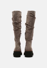 Botas altas de ante color topo con cañas sueltas, cremalleras laterales y tacones gruesos negros, mostradas desde la parte posterior sobre un fondo blanco.