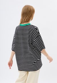 Zwart-wit gestreepte oversized t-shirt met een groene geribbelde halslijn, korte mouwen en een relaxte fit. Witte achtergrond.