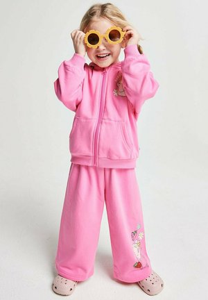 Kleines Kind im pinkfarbenen Trainingsanzug und hellrosa Schuhen, trägt gelbe, blumenförmige Sonnenbrille, lächelt vor einem einfarbigen Hintergrund.