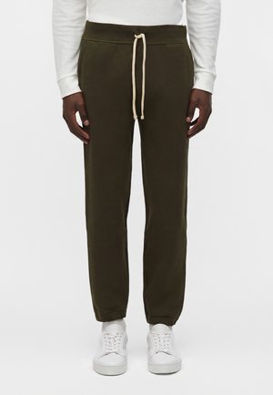 Homme portant un pantalon de jogging vert foncé avec un cordon beige, des baskets blanches et une chemise blanche à manches longues sur un fond uni.