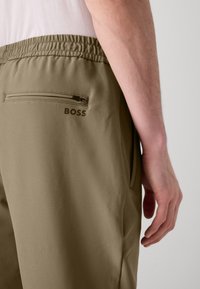 Kaki joggeri ar elastīgu jostasvietu, ar iešūtu kabatu aizmugurē un zīmola logo "BOSS" tumšā burtu drukā.