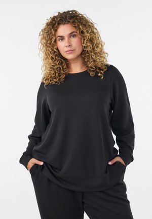 Kobieta o kręconych blond włosach, ubrana w czarną bluzkę z długim rękawem i czarne spodnie, stojąca z rękami w kieszeniach na białym tle.