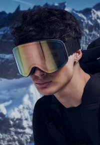 Smučarska očala z veliko, ukrivljeno lečo v prelivnih odtenkih zelene in zlata, obrobljena s črno ter z teksturiranim trakom. V ozadju so zasneženi hribi.