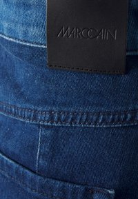 Tmavě modré džínové kalhoty s černou koženou nášivkou na pásu s vyraženým nápisem "MARCCAIN." Jsou viditelné švy a detaily kapes.