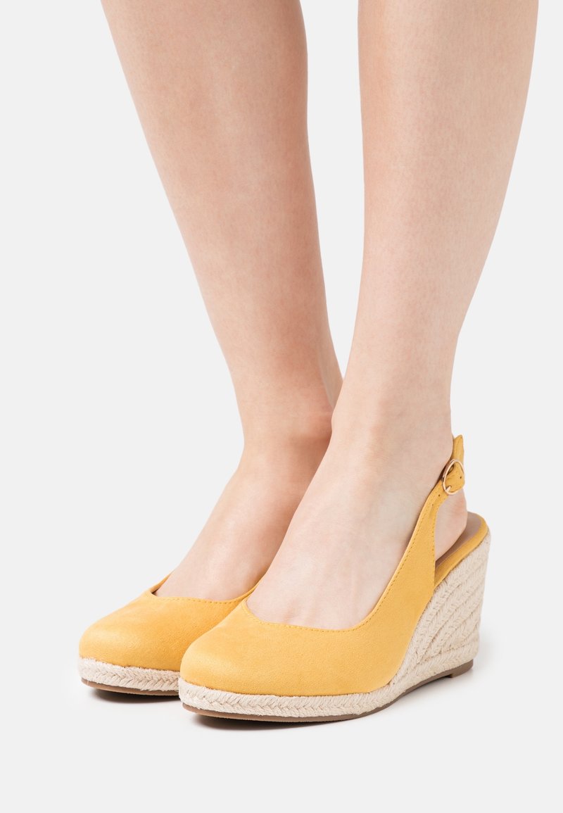 Chaussures compensées à bride arrière en daim jaune aux pieds avec des détails en corde tressée sur les talons compensés sur fond blanc.
