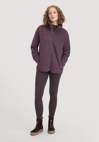 Fleece-tyylinen vetoketjullinen takki tumman violetissa, yhdessä yhteensopivien leggingsien kanssa. Kengissä on tummasta mokasta valmistettu päällinen ja teksturoitu alusta.