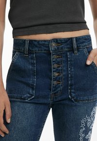 Dark blue denim jeans with a high waist, featuring five metal buttons, angled pockets, and white embroidered butterfly and star details.