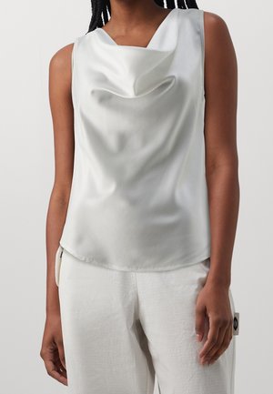 Kvinde iført hvid satin top uden ærmer med cowl hals og hvide teksturerede bukser mod en ensfarvet baggrund.