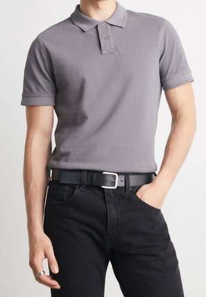 Chemise polo grise avec un col classique, deux boutons, des manches courtes et un logo discret. Associée à un jean noir et une ceinture noire.