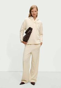 Cream-colored hoodie with drawstrings, paired with wide-leg pants. The model holds a brown shoulder bag and wears pointed brown shoes.