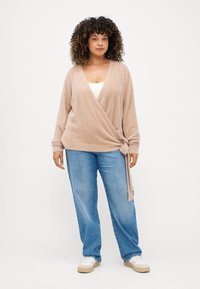 Μπεζ, φουντωτός πλεκτός τύπος κάρντιγκαν με V λαιμόκοψη, μακριά μανίκια και ζωνάκι στη μέση, σε συνδυασμό με ανοιχτό μπλε τζιν straight-leg και αθλητικά παπούτσια.