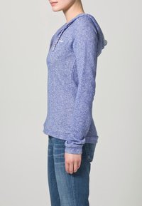 Lila hoodie med melerad färg, dragsko, långa ärmar och en avslappnad passform, kombinerad med blå denimjeans. Mjuk textur och avslappnad design.