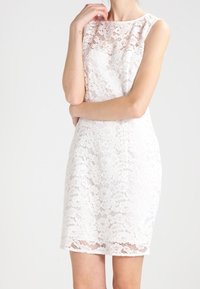 Sukienka koronkowa w kolorze białym o dopasowanym kroju, z dekoltem karo i bezrękawnikowym fasonem. Charakteryzuje się kwiatowymi wzorami koronkowymi i prostym wykończeniem dołu.