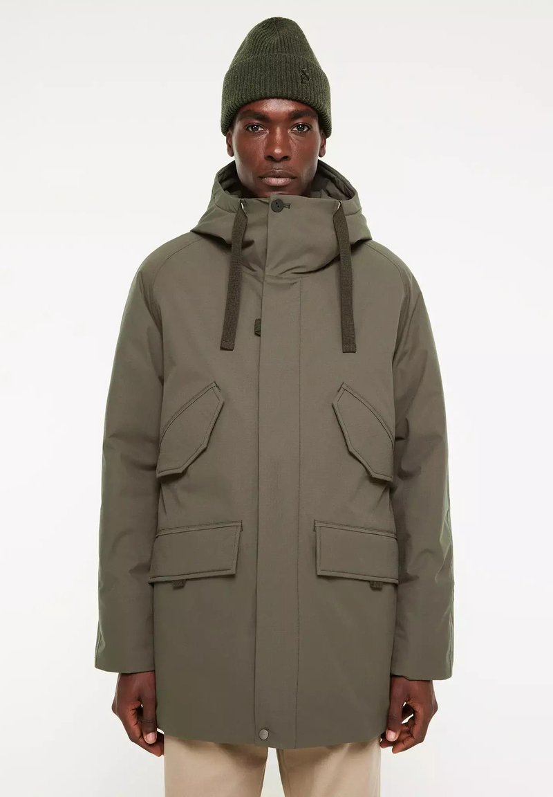 Olijfgroene parka met een hoge kraag, verstelbare capuchon, meerdere voorkruiden en een gladde textuur, gedragen over lichtgekleurde broek.